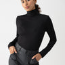 A person with dark, curly hair pulled back is wearing the Essential Turtleneck Top Black by Sanctuary Clothing and black high-waisted pants. They stand with hands in pockets, looking confidently at the camera against a plain light background.