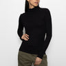 A person with long dark hair wears the Sanctuary Clothing essential turtleneck top in black and olive green pants, posing against a plain white background. They look to the side with one hand on their hip.