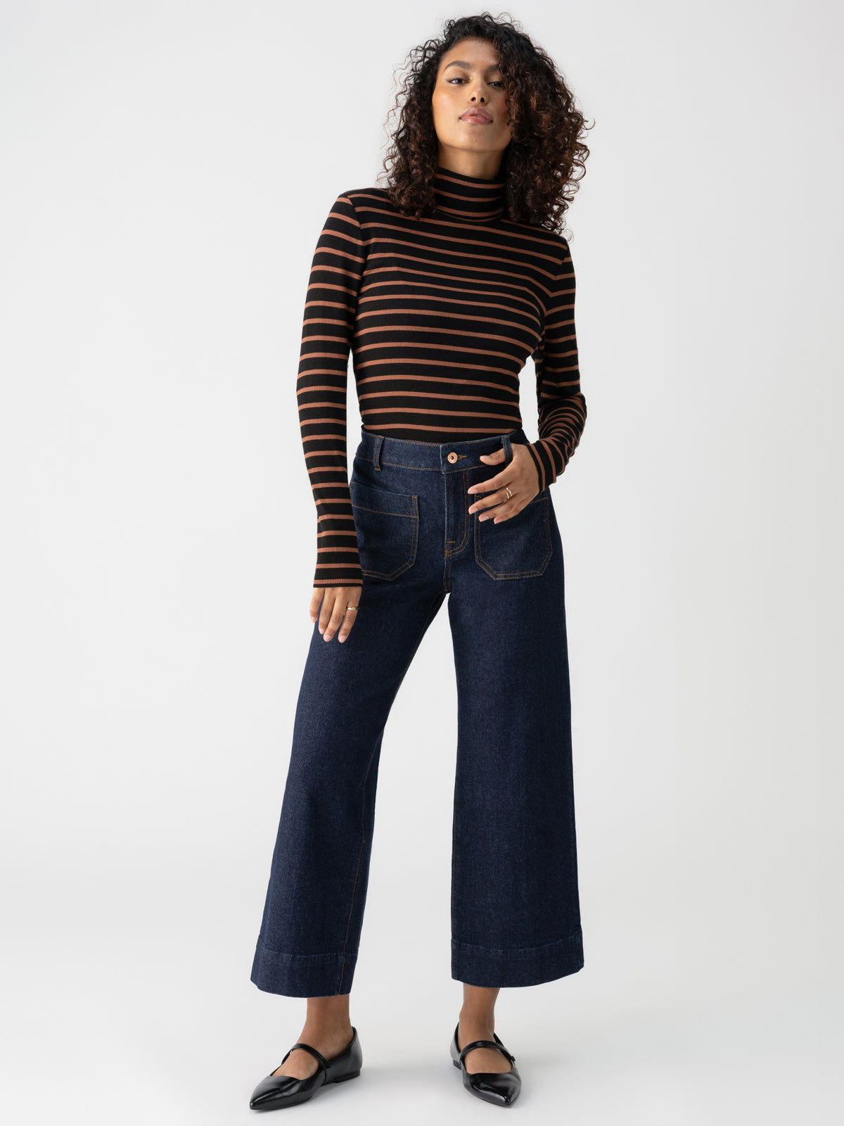 A woman with curly hair stands against a plain background, wearing the Essential Turtleneck Raw Hide Black Stripe by Sanctuary Clothing, wide-leg dark blue jeans with front pockets, and black flats. Her left hand rests in her pocket while her right arm hangs by her side.