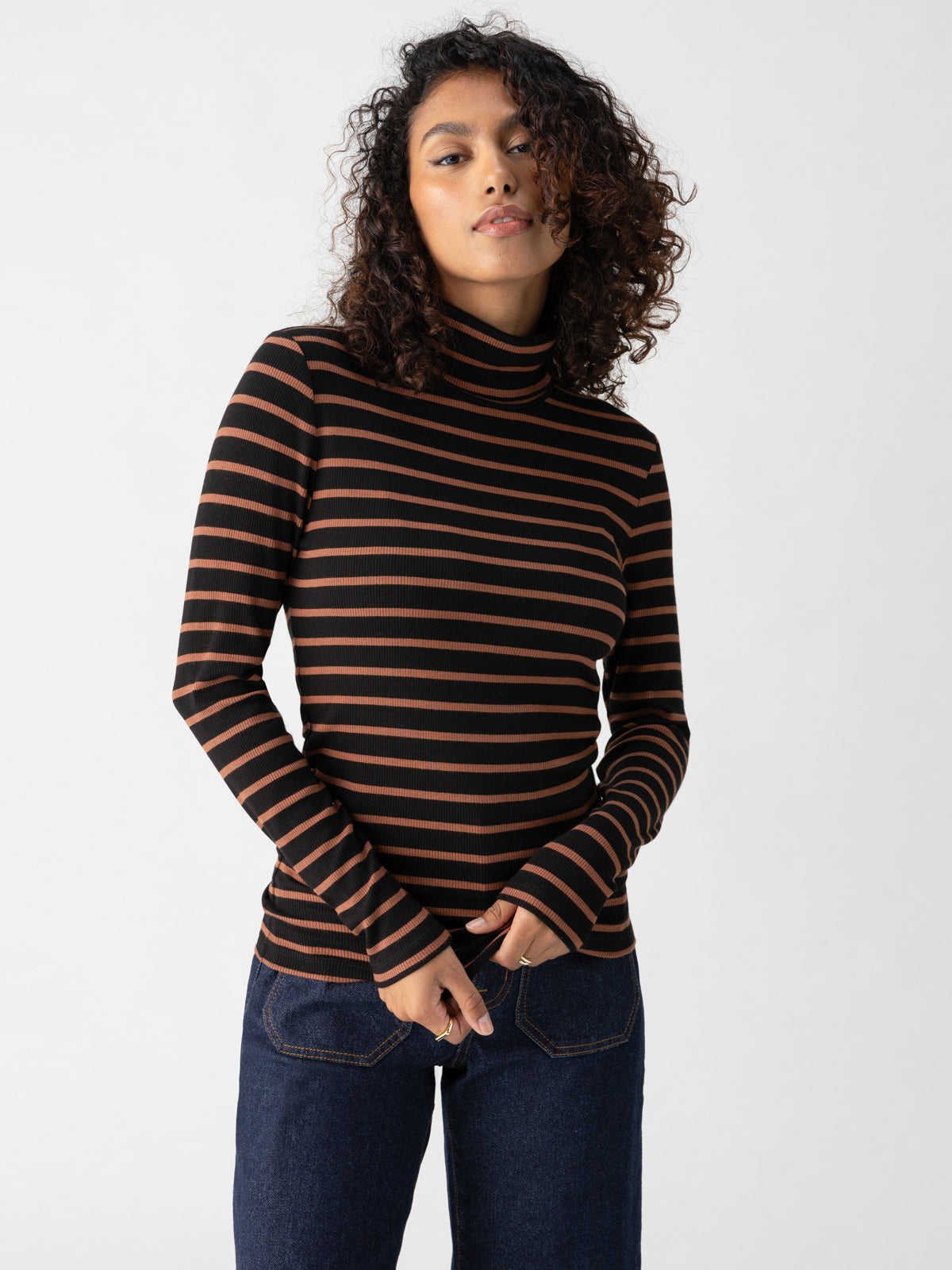 A person with curly hair, wearing the Essential Turtleneck Raw Hide Black Stripe by Sanctuary Clothing and dark blue jeans, stands against a plain white background, looking confidently at the camera with hands loosely clasped in front.