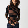 A person with curly hair, wearing the Essential Turtleneck Raw Hide Black Stripe by Sanctuary Clothing and dark blue jeans, stands against a plain white background, looking confidently at the camera with hands loosely clasped in front.