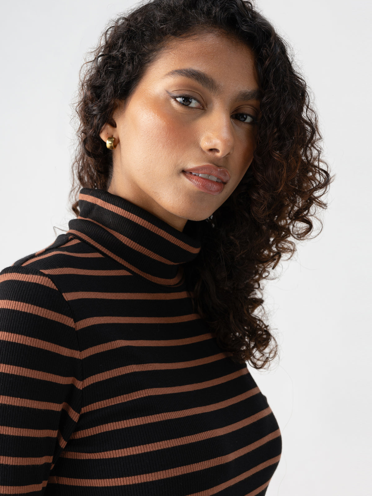 A person with medium-length curly hair wears the Essential Turtleneck Raw Hide Black Stripe from Sanctuary Clothing. They have small gold earrings and are turned slightly to the side, looking at the camera with a neutral expression. The background is plain white.