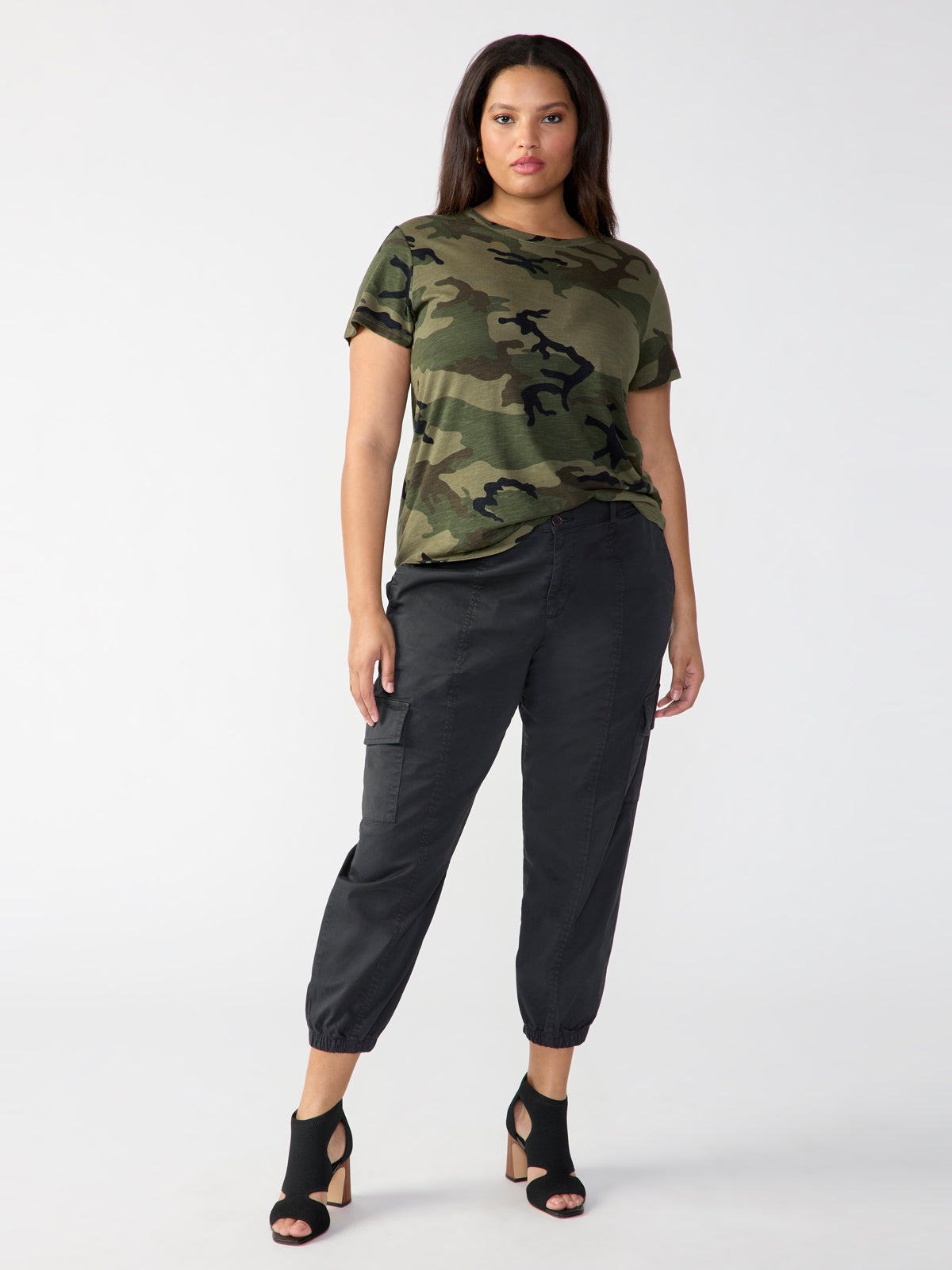 A person stands confidently against a plain background, wearing "The Perfect Tee Hiker Camo Inclusive Collection" by Sanctuary Clothing and black cargo pants. They have black open-toe heeled shoes. Their hands are relaxed by their sides.