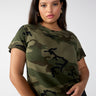 A person with long hair is wearing "The Perfect Tee Hiker Camo Inclusive Collection" by Sanctuary Clothing and black pants. They have gold hoop earrings and are standing against a plain light grey background. Their expression is neutral, and they are looking directly at the camera with one hand resting on their stomach.