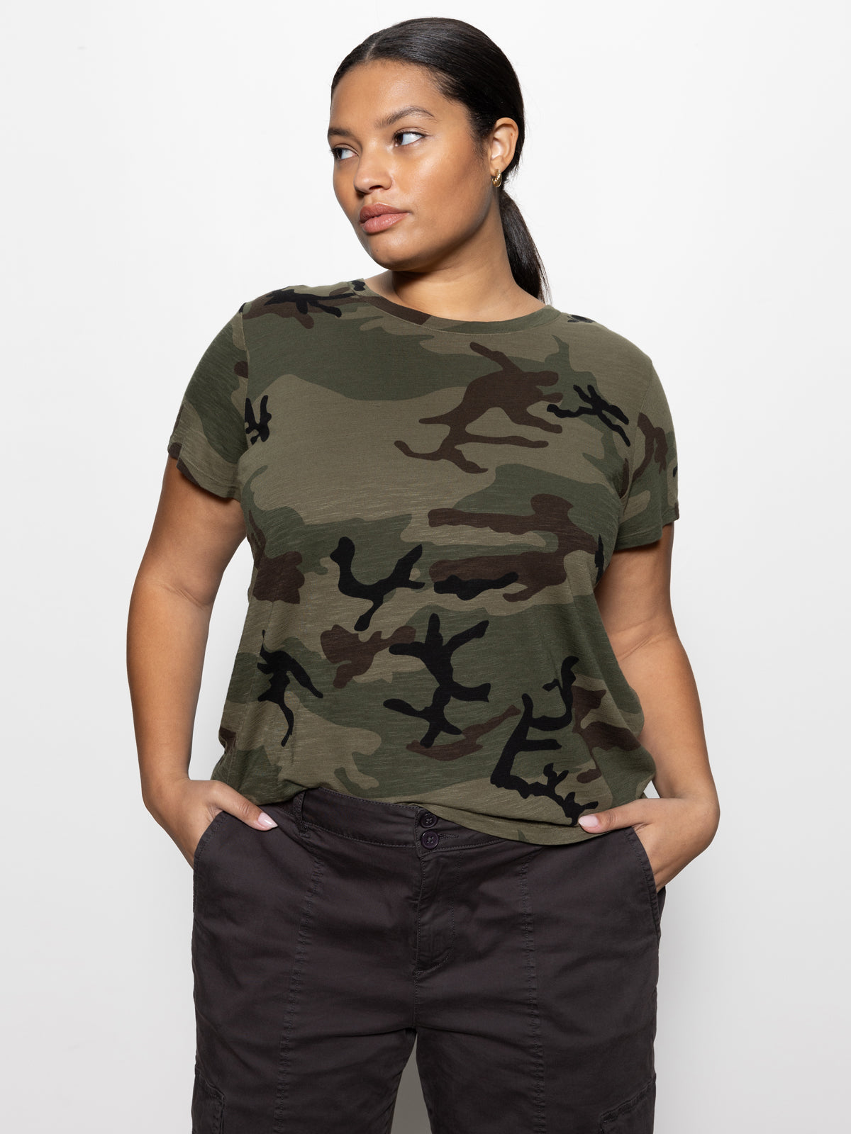 A person in the Sanctuary Clothing "Perfect Tee" from the Hiker Camo Inclusive Collection and dark pants stands against a white background, with hands in pockets. They have long, dark hair pulled back and gaze to the side.