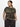 A person in the Sanctuary Clothing "Perfect Tee" from the Hiker Camo Inclusive Collection and dark pants stands against a white background, with hands in pockets. They have long, dark hair pulled back and gaze to the side.