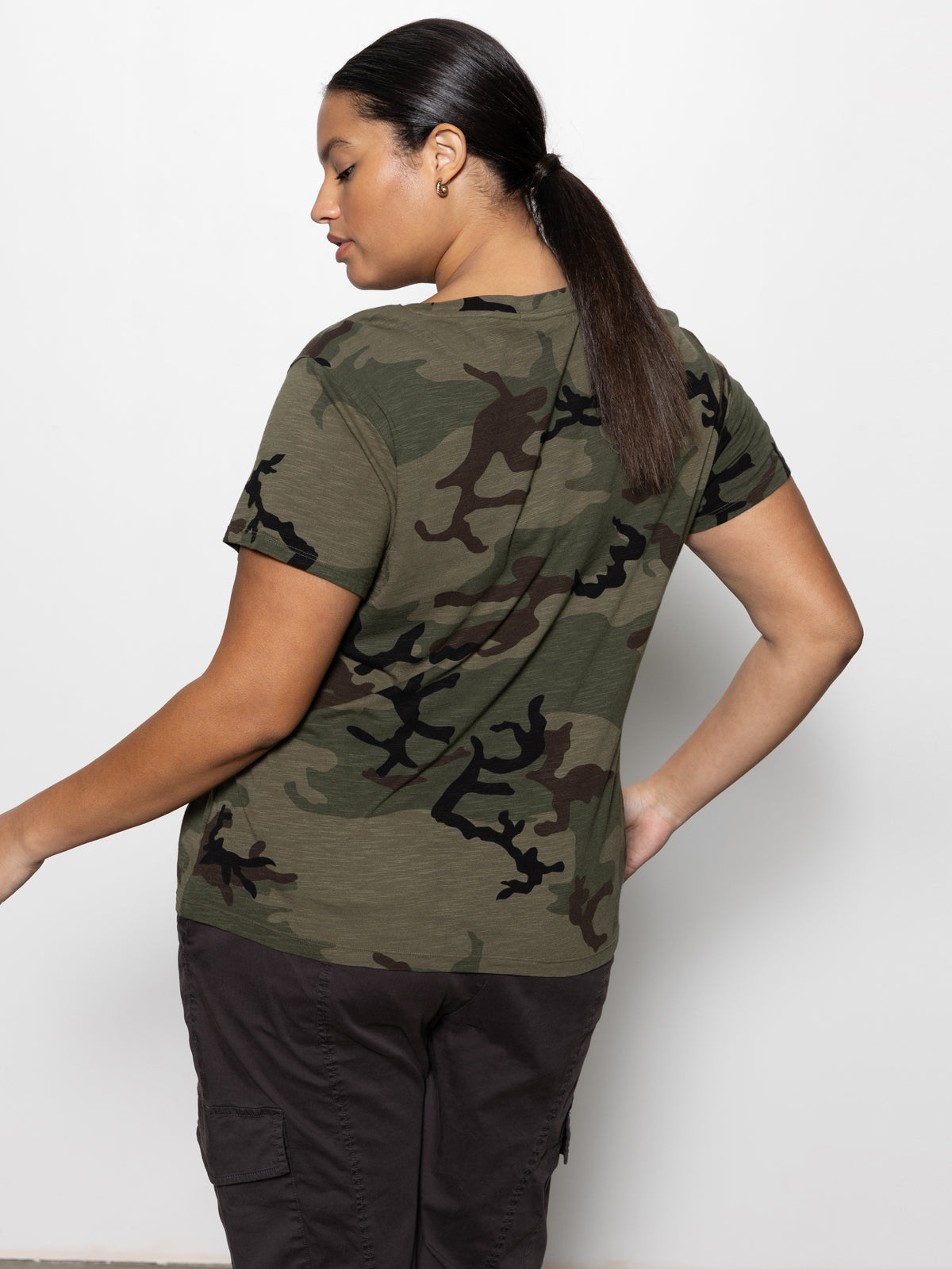 A person models the perfect tee hiker camo inclusive collection by Sanctuary Clothing, showcasing its green and brown camouflage design. They have long dark hair tied back and are paired with dark cargo pants against a plain white background.