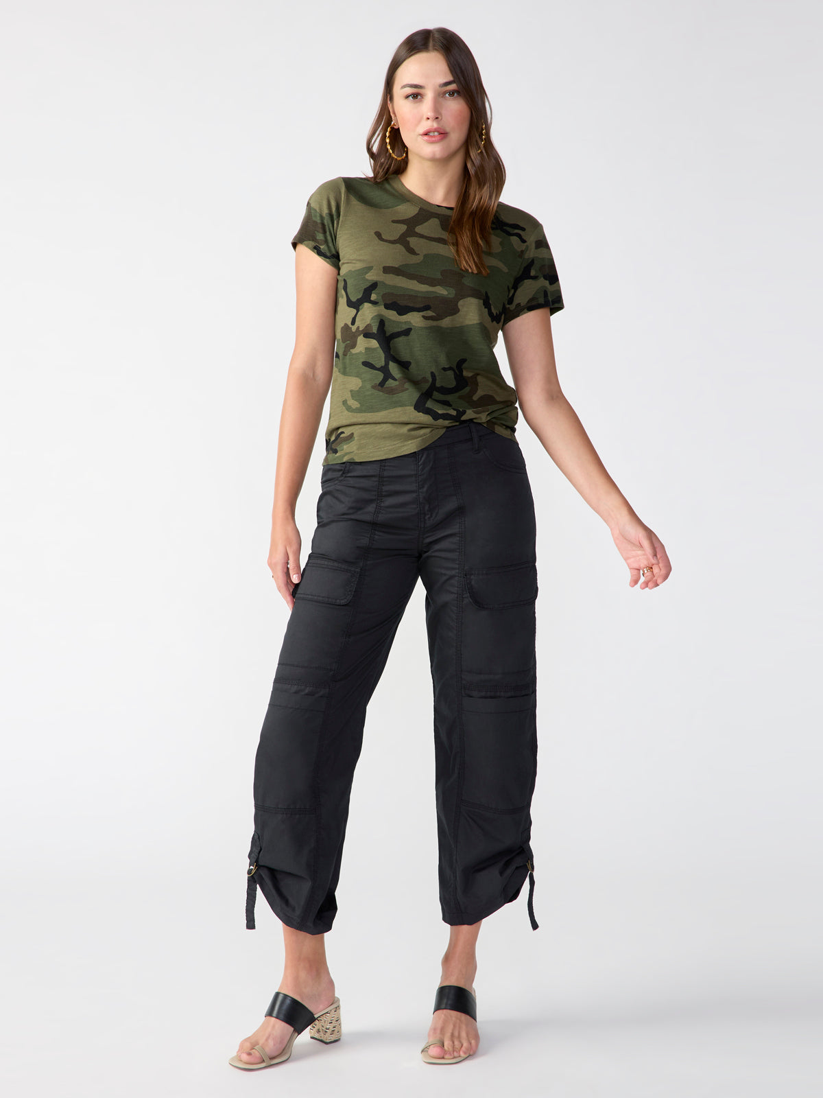 A woman stands against a plain background wearing "The Perfect Tee Hiker Camo" by Sanctuary Clothing paired with black cargo pants featuring drawstring hems. She has long hair and large hoop earrings, and she's wearing black sandals with a low heel. One of her hands rests at her side while the other is slightly raised.