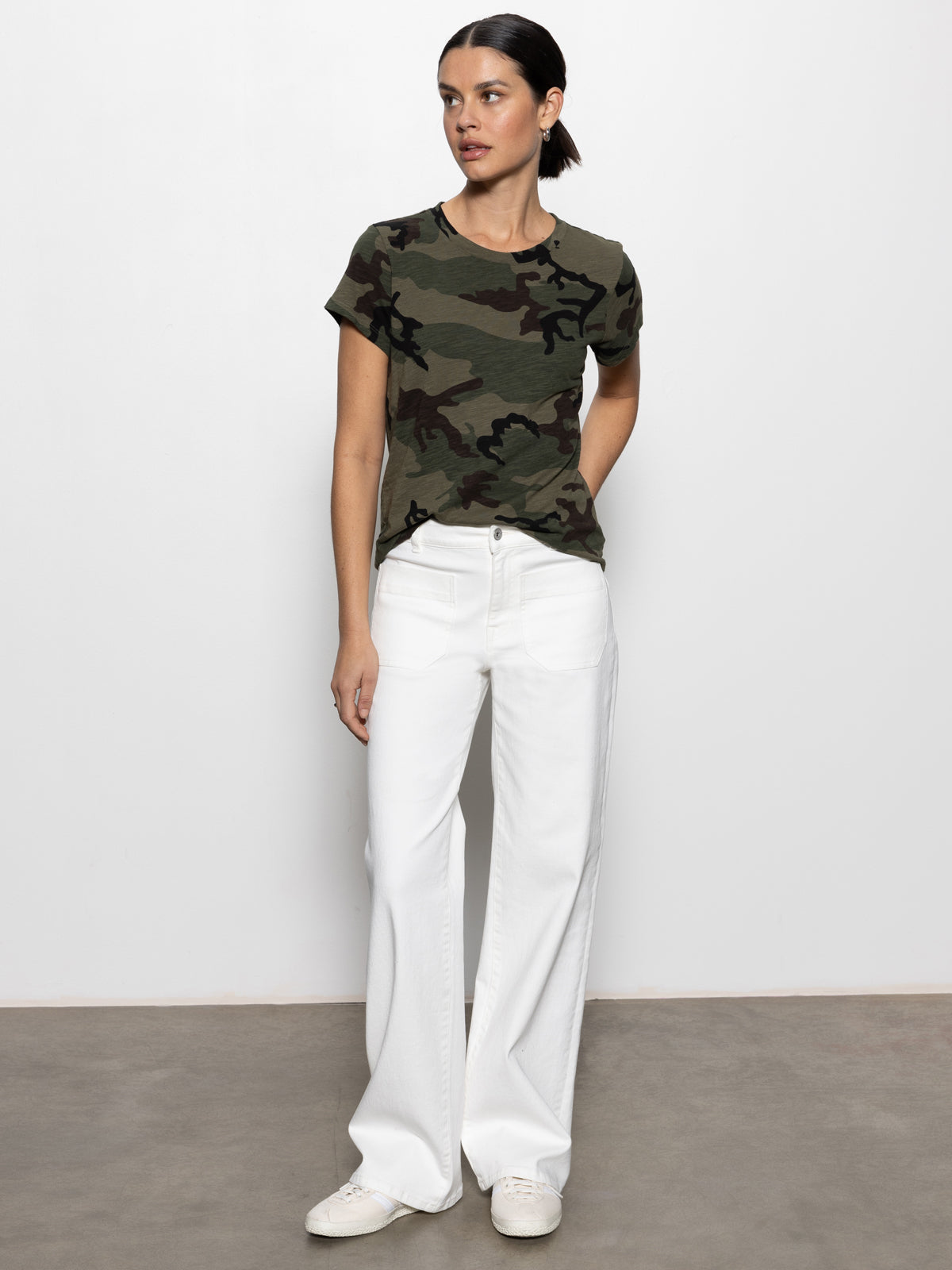 Against a plain background, a person wears "the perfect tee hiker camo" by Sanctuary Clothing with white wide-leg pants. They have dark hair tied back and look to the side, standing on a simple concrete floor.