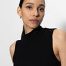 A woman with long dark hair tied back is wearing the Sanctuary Clothing essential sleeveless mock neck top in black. She has gold hoop earrings, a neutral expression, and is set against a plain light background while looking into the camera.