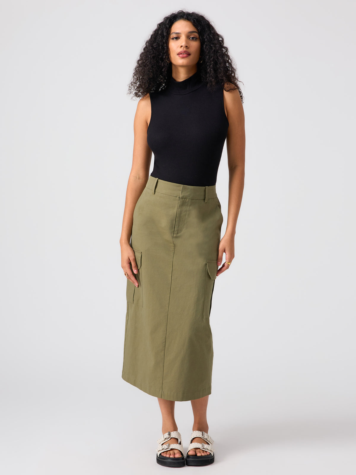 A woman with curly hair is wearing a black Essential Sleeveless Mock Neck Top by Sanctuary Clothing, paired with an olive green midi skirt featuring cargo pockets. She stands facing forward with her hands relaxed by her sides, and completes her outfit with white sandals that have thick straps.