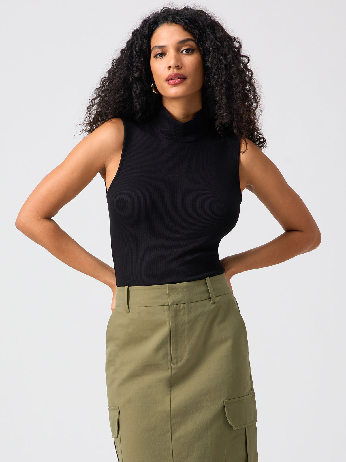 A woman with long, curly hair is wearing the Essential Sleeveless Mock Neck Top in black by Sanctuary Clothing, tucked into a high-waisted olive green skirt with pockets. She is standing against a plain, light gray background. Her hands rest on her hips and she looks directly at the camera.