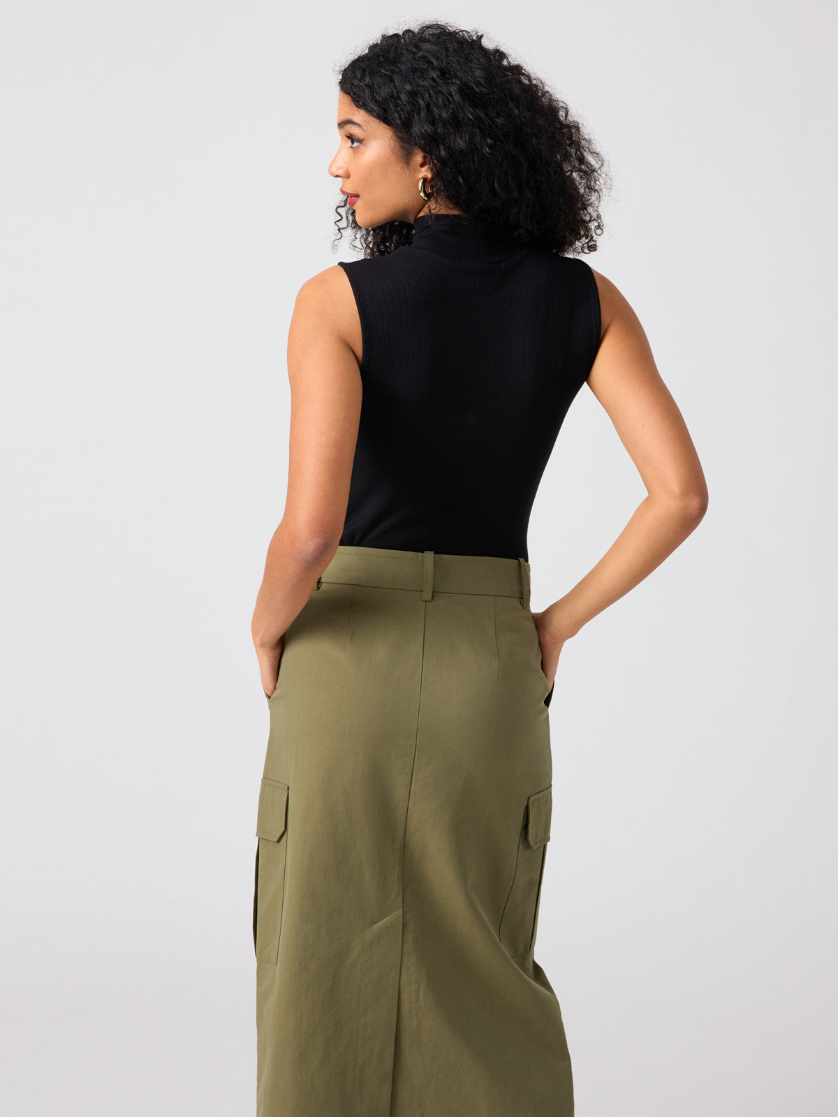 A person with curly hair is standing and facing away from the camera. They are wearing the Essential Sleeveless Mock Neck Top in black by Sanctuary Clothing and an olive green, high-waisted cargo skirt with their hands in the skirt's pockets. The background is plain and light-colored.