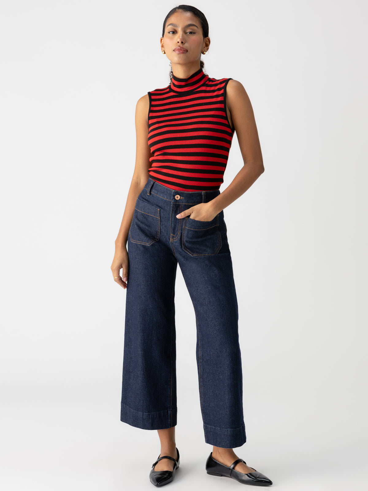 A person stands against a plain white background, wearing the Essential Sleeveless Mock Neck in Black Cherry Red Stripe by Sanctuary Clothing, high-waisted dark blue wide-leg jeans with front pockets, and black ballet flats. One hand is in their pocket while the other hangs by their side.