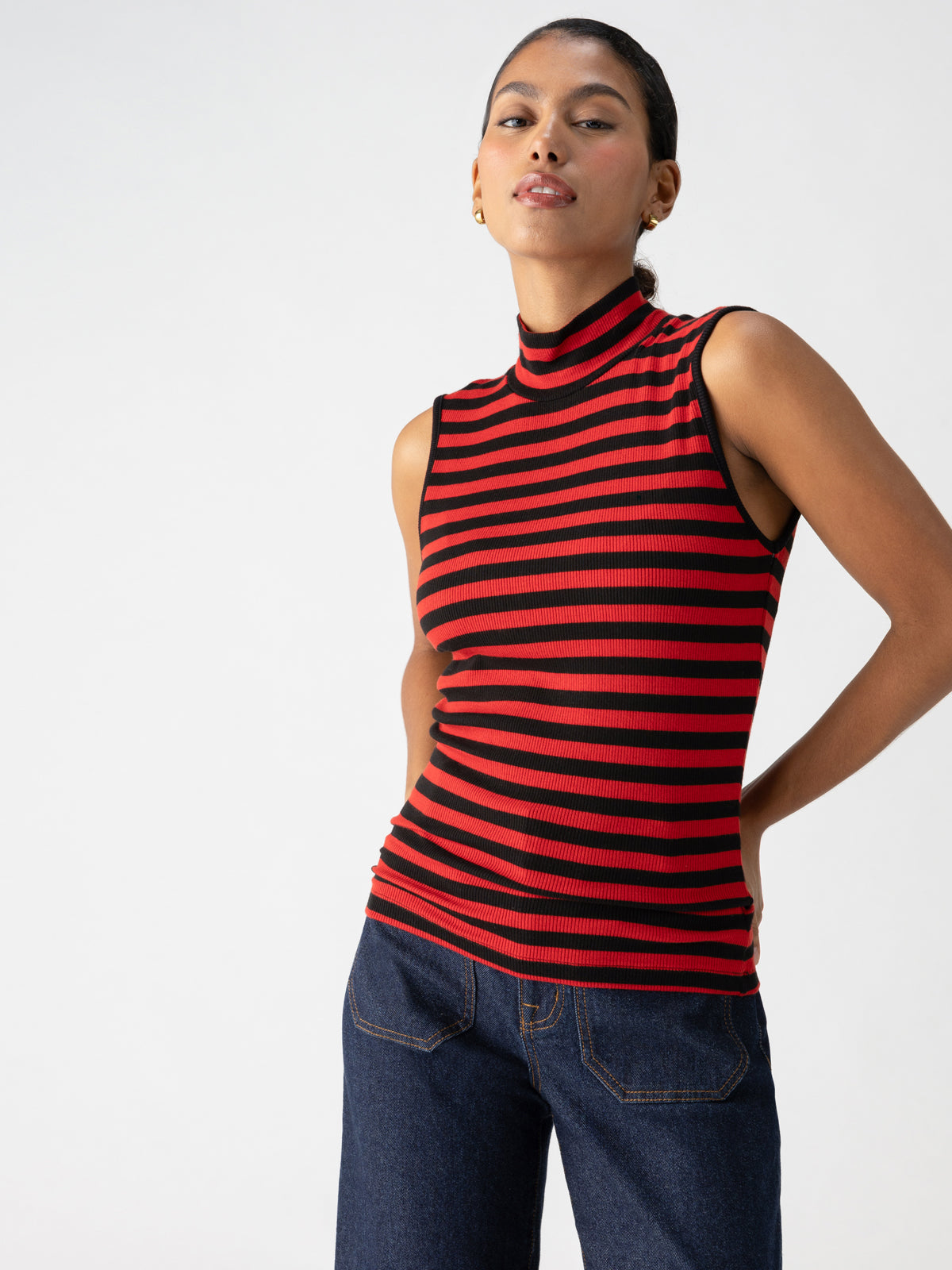 A person stands confidently against a plain background, wearing the Essential Sleeveless Mock Neck Black Cherry Red Stripe top from Sanctuary Clothing and dark blue jeans. They have one hand on their hip, and their hair is pulled back. The expression on their face is neutral.