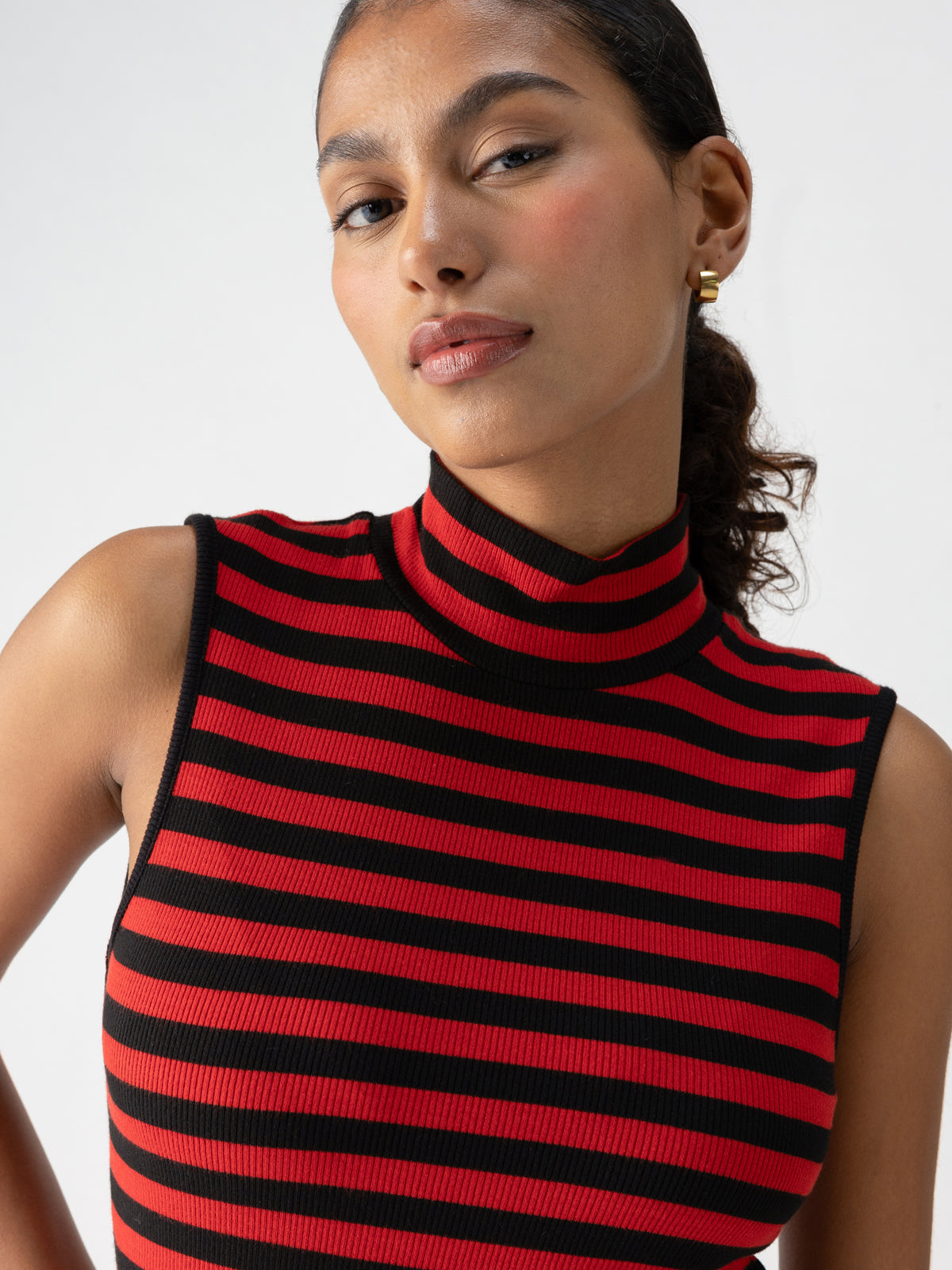 A person with pulled-back hair is wearing the "Essential Sleeveless Mock Neck Black Cherry Red Stripe" top by Sanctuary Clothing. They are looking directly at the camera with a neutral expression, and gold hoop earrings are visible on their ears. The background is plain white.