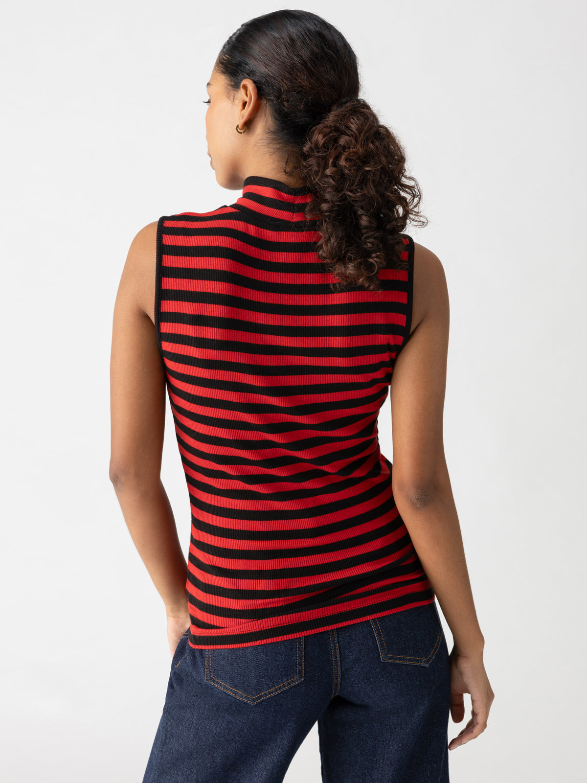 A person with curly hair tied back into a ponytail is standing with their back to the camera. They are wearing the Sanctuary Clothing Essential Sleeveless Mock Neck top in Black Cherry Red Stripe, paired with dark blue jeans. The background is plain and white.