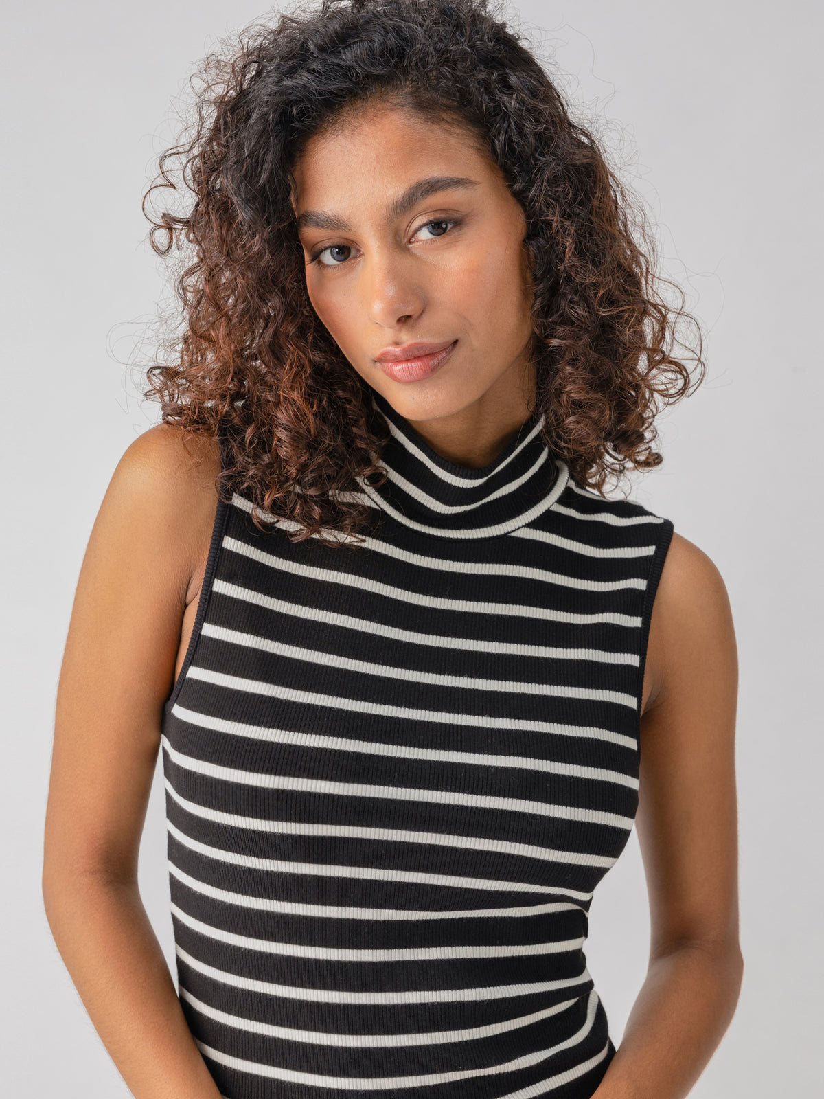 A person with curly hair is wearing the Essential Sleeveless Mock Neck Black Chalk Stripe top from Sanctuary Clothing. They are standing against a plain, light grey background and gazing towards the camera with a neutral expression.