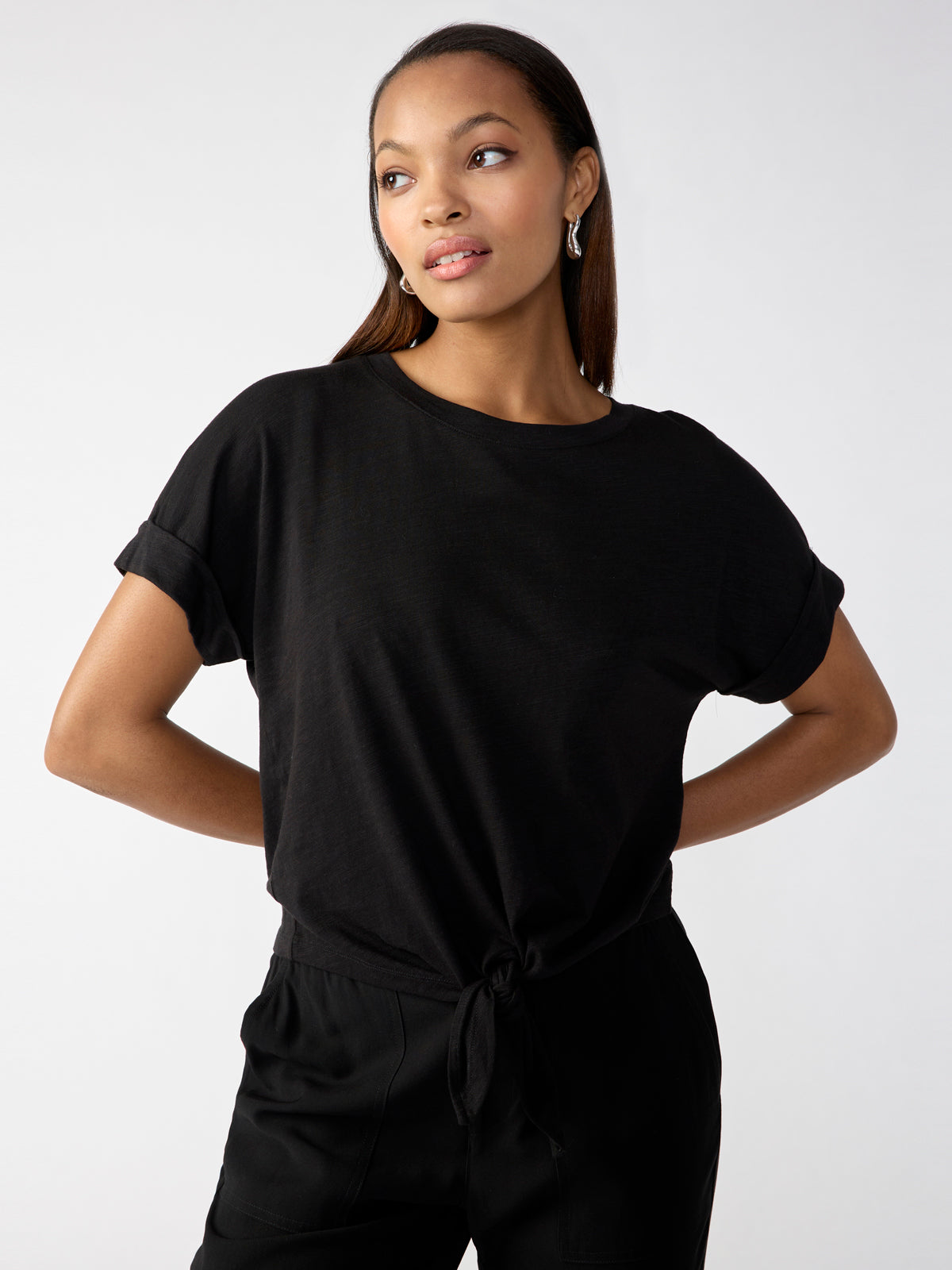 A woman poses confidently against a plain background. She wears the All Day Tie Tee Black from Sanctuary Clothing's Inclusive Collection, paired with black pants. Her straight hair is parted to one side, and she accessorizes with hoop earrings. With her hands resting on her hips, she gazes slightly to the side.