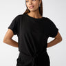 A woman poses confidently against a plain background. She wears the All Day Tie Tee Black from Sanctuary Clothing's Inclusive Collection, paired with black pants. Her straight hair is parted to one side, and she accessorizes with hoop earrings. With her hands resting on her hips, she gazes slightly to the side.