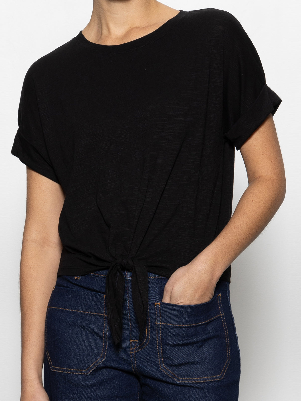 A person wearing Sanctuary Clothing's all day tie tee black from the inclusive collection, paired with blue jeans, has one hand in their pocket and is standing against a plain white background.