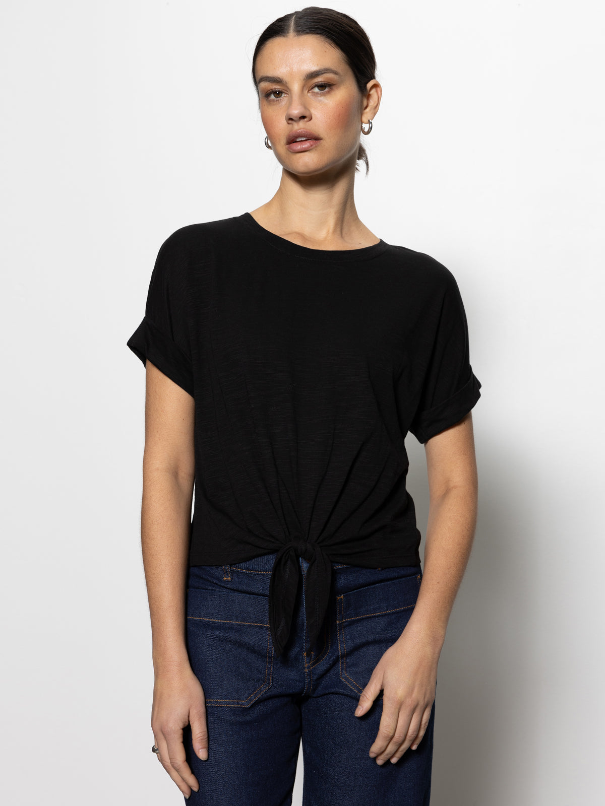 A person models the all day tie tee black from Sanctuary Clothing's inclusive collection, paired with dark jeans. They have dark hair tied back and wear small hoop earrings, posing against a plain white background.