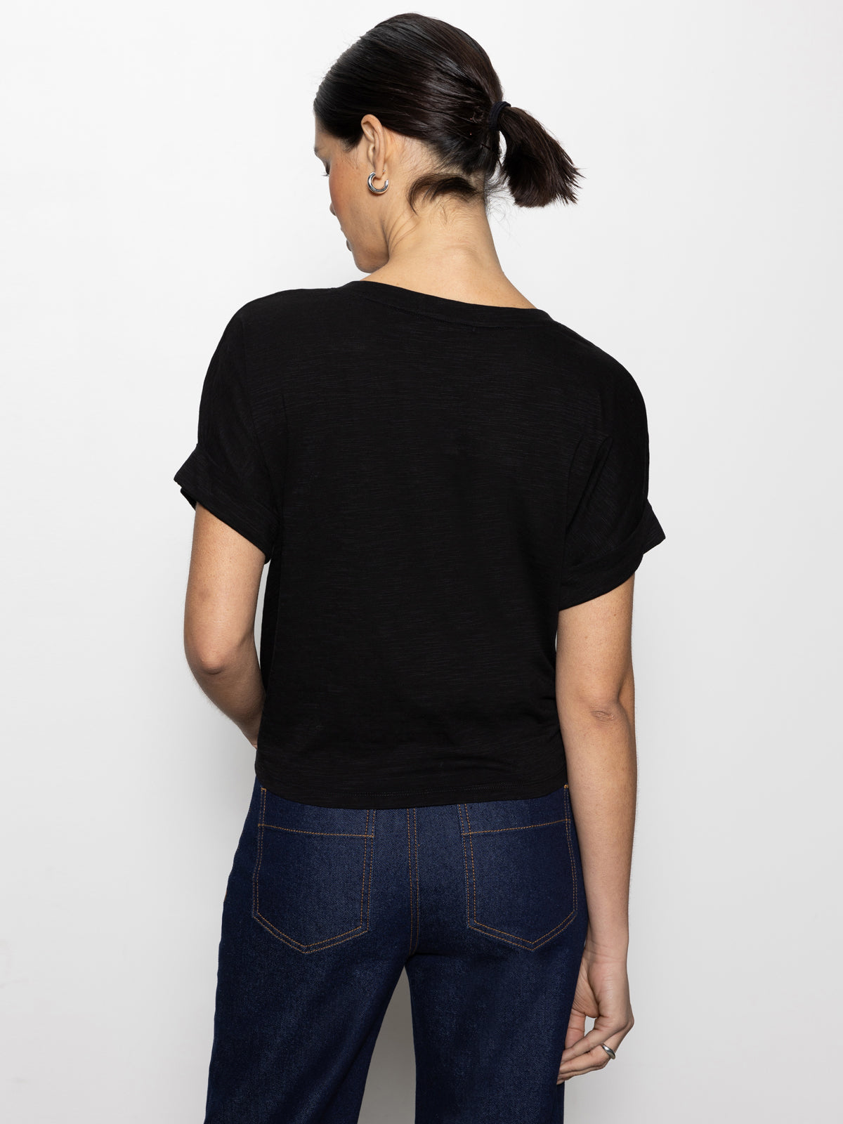 A person with dark hair tied back is facing away, wearing Sanctuary Clothing's 'all day tie tee black inclusive collection' and dark blue jeans with visible pockets against a plain white background.