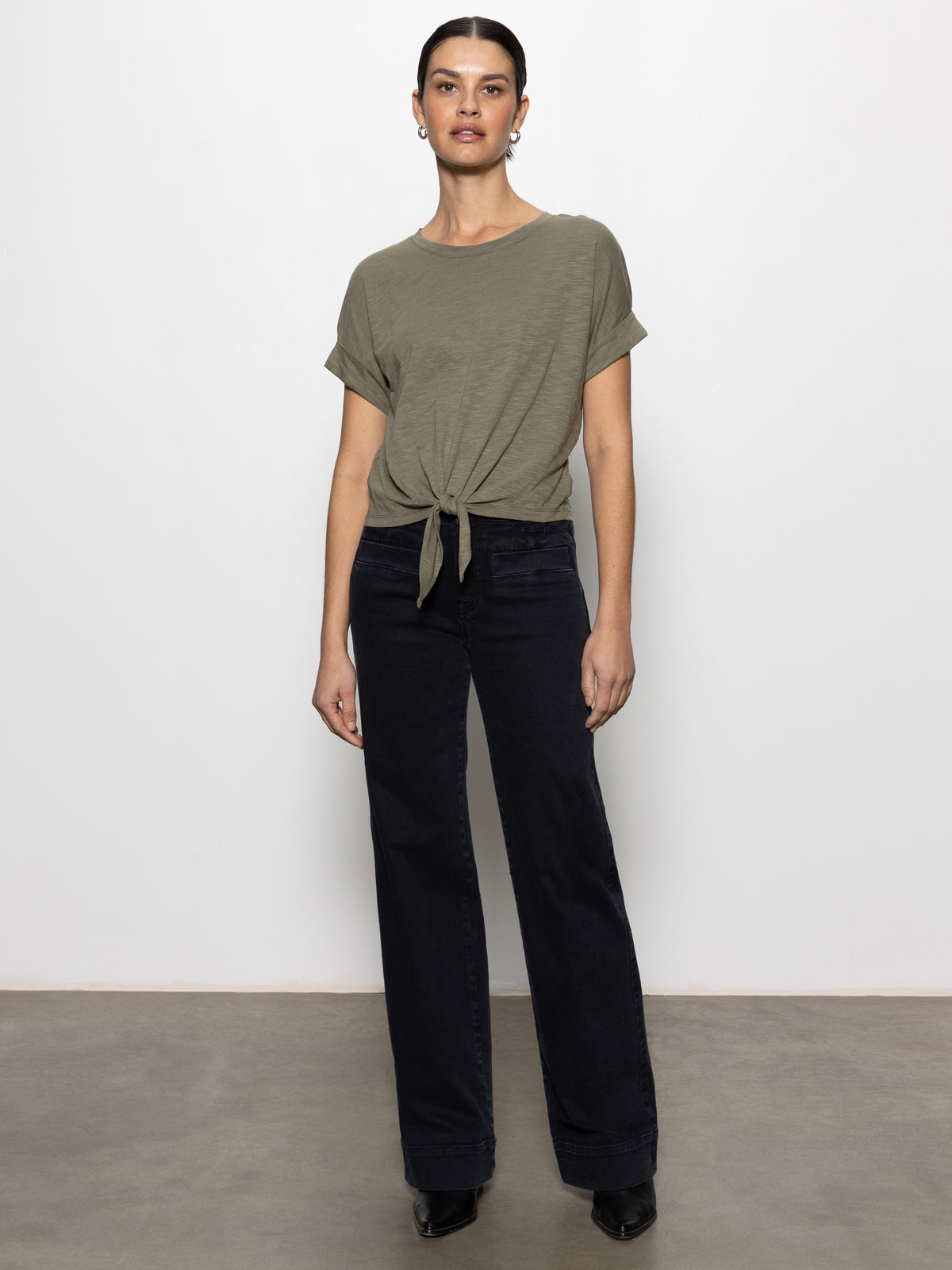 A person stands by a plain white wall, dressed in the Sanctuary Clothing all day tie tee trail green, paired with dark wide-leg pants and black shoes. Their hair is tied back and their arms are relaxed by their sides.