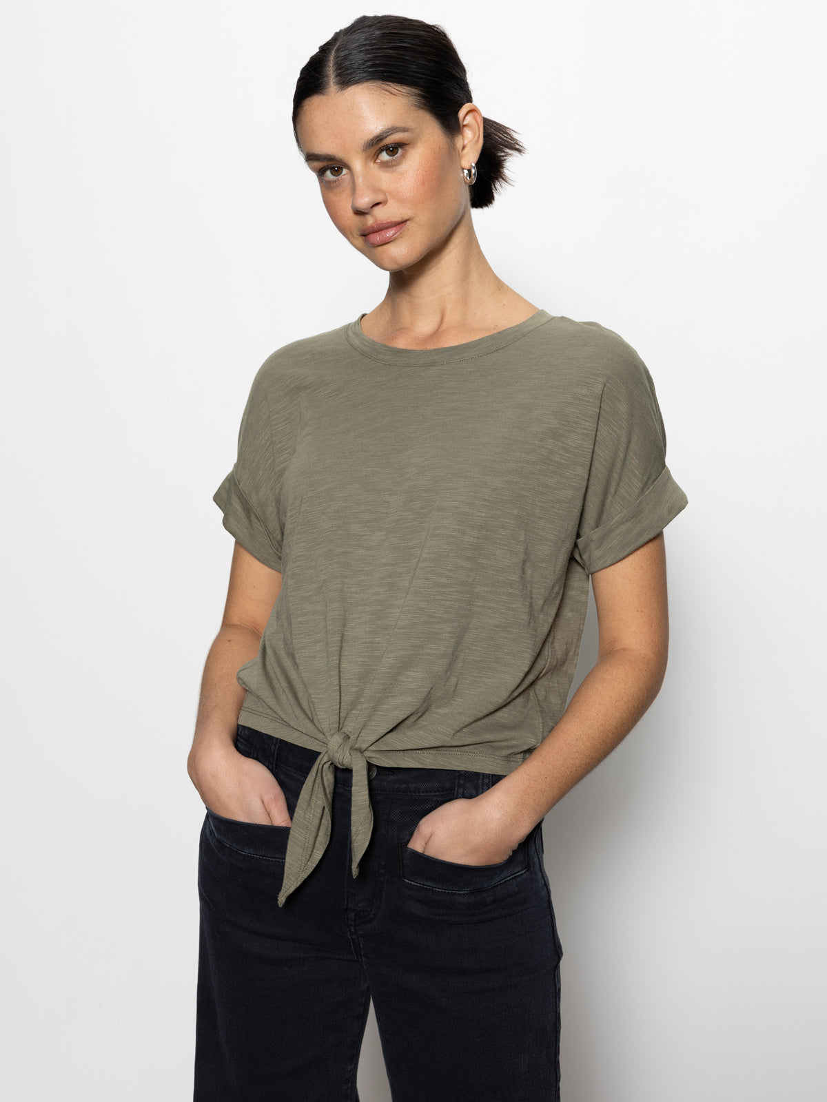 A person with dark hair tied back is wearing the Sanctuary Clothing all day tie tee in trail green paired with dark jeans. They stand against a plain white backdrop, hands in pockets, exuding confidence as they look at the camera.