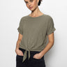 A person with dark hair tied back is wearing the Sanctuary Clothing all day tie tee in trail green paired with dark jeans. They stand against a plain white backdrop, hands in pockets, exuding confidence as they look at the camera.