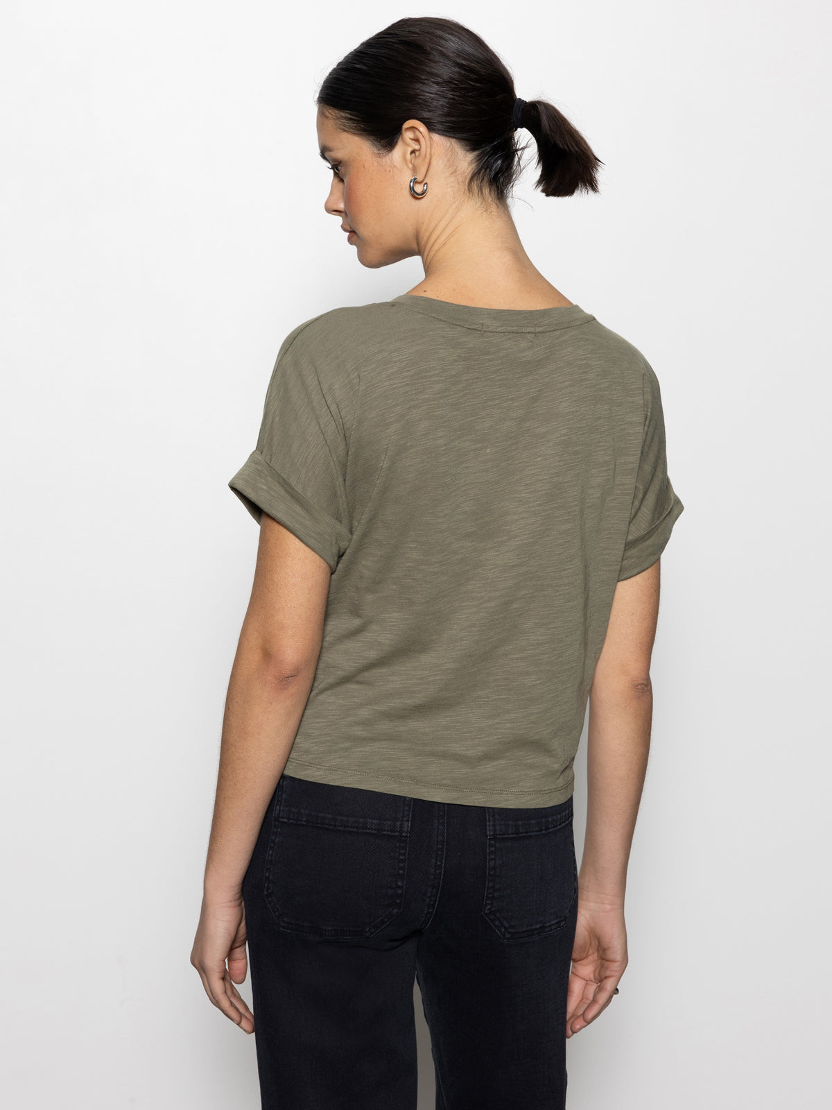 A woman with dark hair in a low ponytail faces sideways, wearing Sanctuary Clothing's "all day tie tee" in trail green and dark jeans against a plain white background.