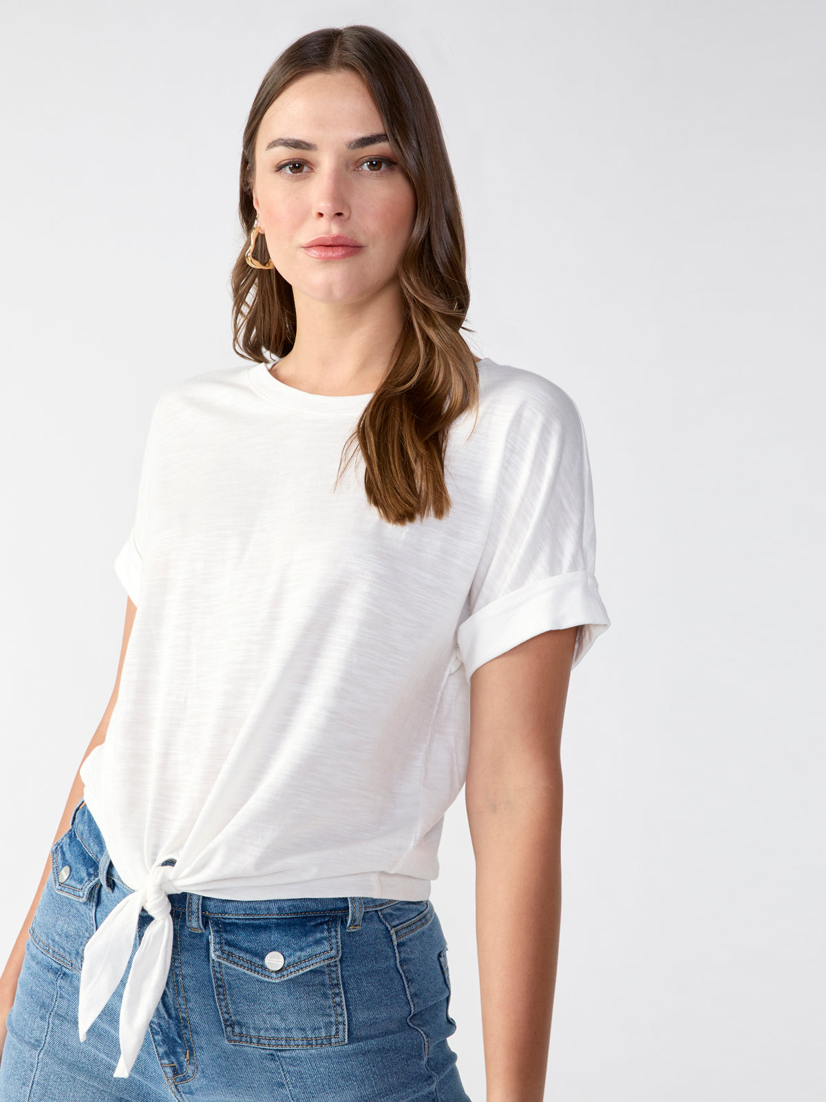 A woman with long brown hair is wearing the Sanctuary Clothing All Day Tie Tee White from the Inclusive Collection and high-waisted blue jeans. She stands facing the camera with a neutral expression against a plain, light-colored background. Her look is complemented by gold hoop earrings.