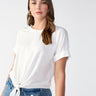 A woman with long brown hair is wearing the Sanctuary Clothing All Day Tie Tee White from the Inclusive Collection and high-waisted blue jeans. She stands facing the camera with a neutral expression against a plain, light-colored background. Her look is complemented by gold hoop earrings.