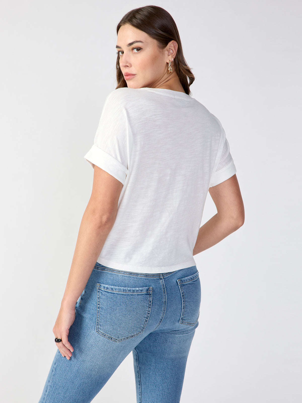 A woman with long brown hair is posing with her back turned towards the camera, looking over her shoulder. She is wearing a white All Day Tie Tee from Sanctuary Clothing's Inclusive Collection and high-waisted blue jeans. The background is plain and light-colored.