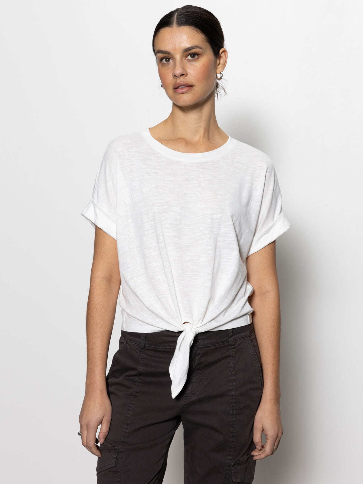 A person stands against a plain white background wearing Sanctuary Clothing's All Day Tie Tee from the Inclusive Collection. The white T-shirt features a front knot and short sleeves, paired with black pants. The person has dark hair pulled back.