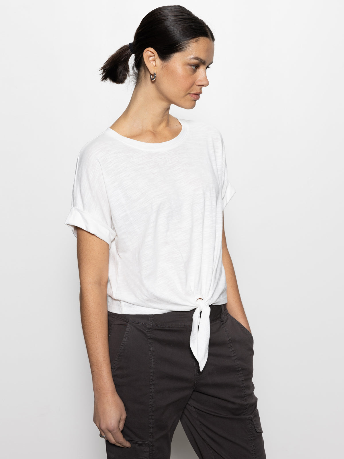 A person with dark hair in a ponytail wears the Sanctuary Clothing "All Day Tie Tee White" from the Inclusive Collection, paired with dark pants. They are gazing sideways against a plain white backdrop.