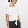 A person with dark hair in a ponytail wears the Sanctuary Clothing "All Day Tie Tee White" from the Inclusive Collection, paired with dark pants. They are gazing sideways against a plain white backdrop.