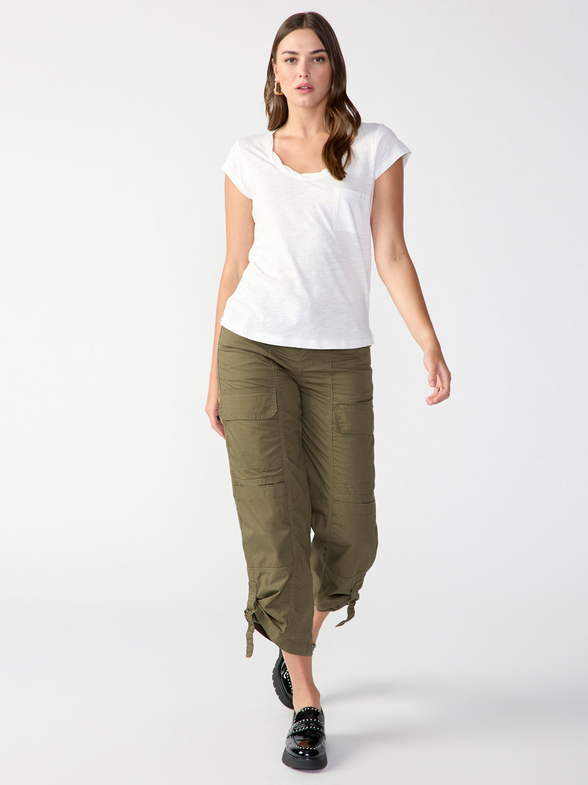 A woman is standing against a plain white background. She is dressed in a casual outfit featuring the "Traveler Twist Tee White" from Sanctuary Clothing, paired with olive-green cargo pants that have drawstring cuffs. She has long brown hair and is wearing black studded shoes.