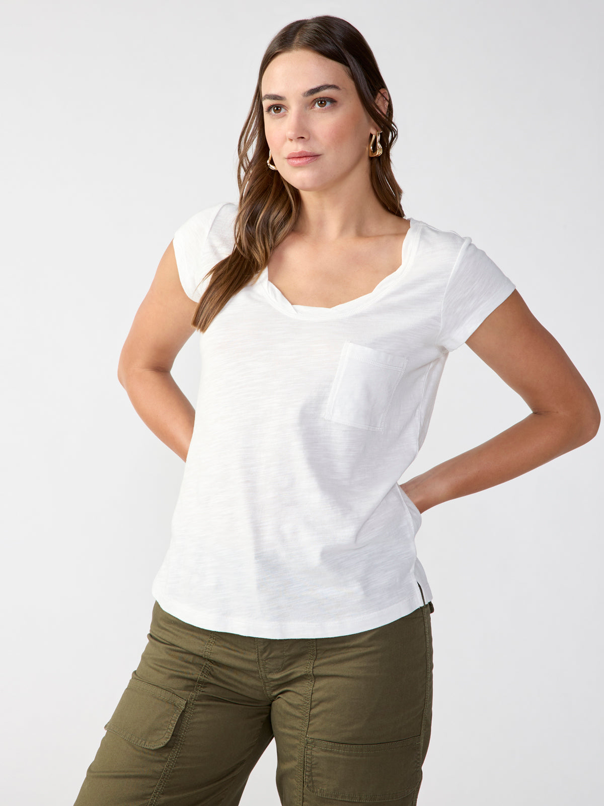 A woman with long, straight hair, partially tied back, stands against a plain background. She is wearing the Traveler Twist Tee White by Sanctuary Clothing and olive green cargo pants. Her hands are positioned on her hips.