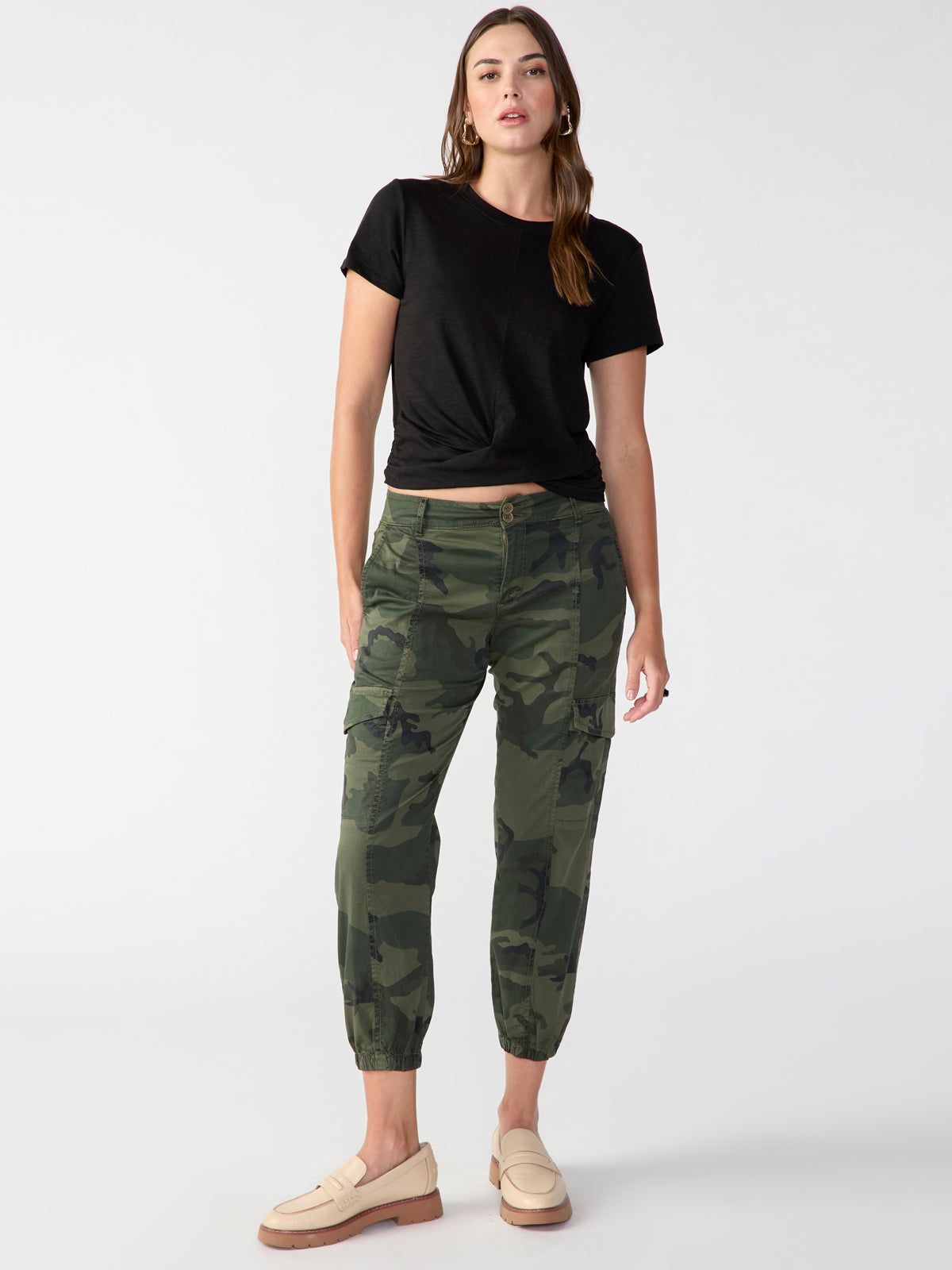 A woman stands confidently against a plain background, wearing the Highstreet Tee Black from Sanctuary Clothing and green camouflage pants. She has light brown hair and is accessorized with hoop earrings. She completes her outfit with beige loafers.