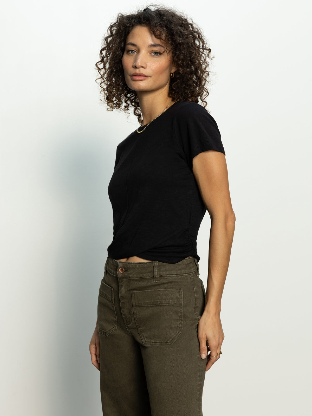 Wearing Sanctuary Clothing's highstreet tee in black and olive green pants, a person with curly hair stands against a plain background, gazing at the camera with a neutral expression.