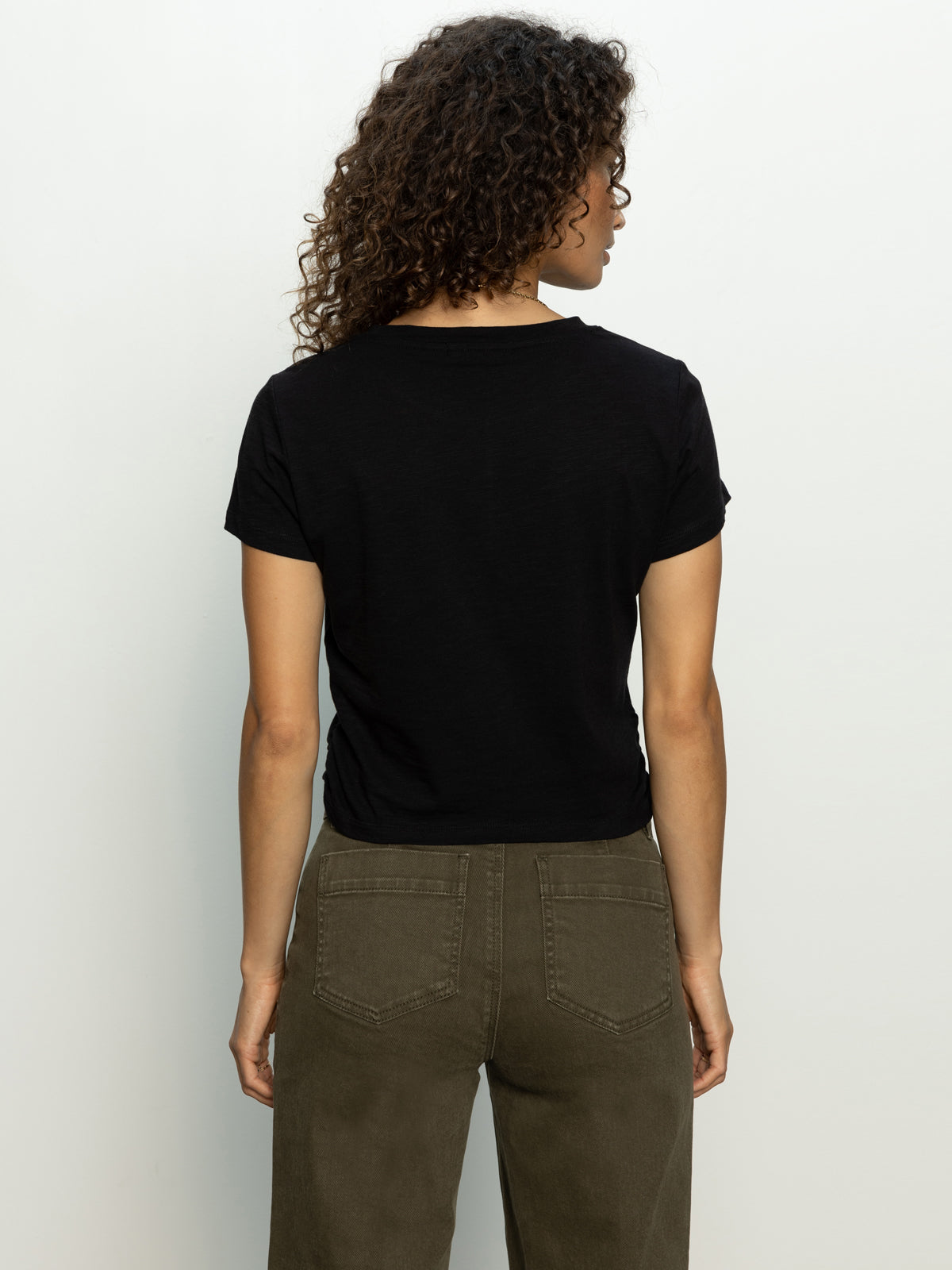 Wearing a highstreet tee in black by Sanctuary Clothing and olive green pants, a person with curly hair stands against a plain light background, facing away.