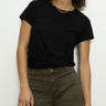 A person wearing Sanctuary Clothing's highstreet tee black and olive green pants stands against a plain white background. They have curly hair, relaxed hands by their sides, and are wearing a necklace.