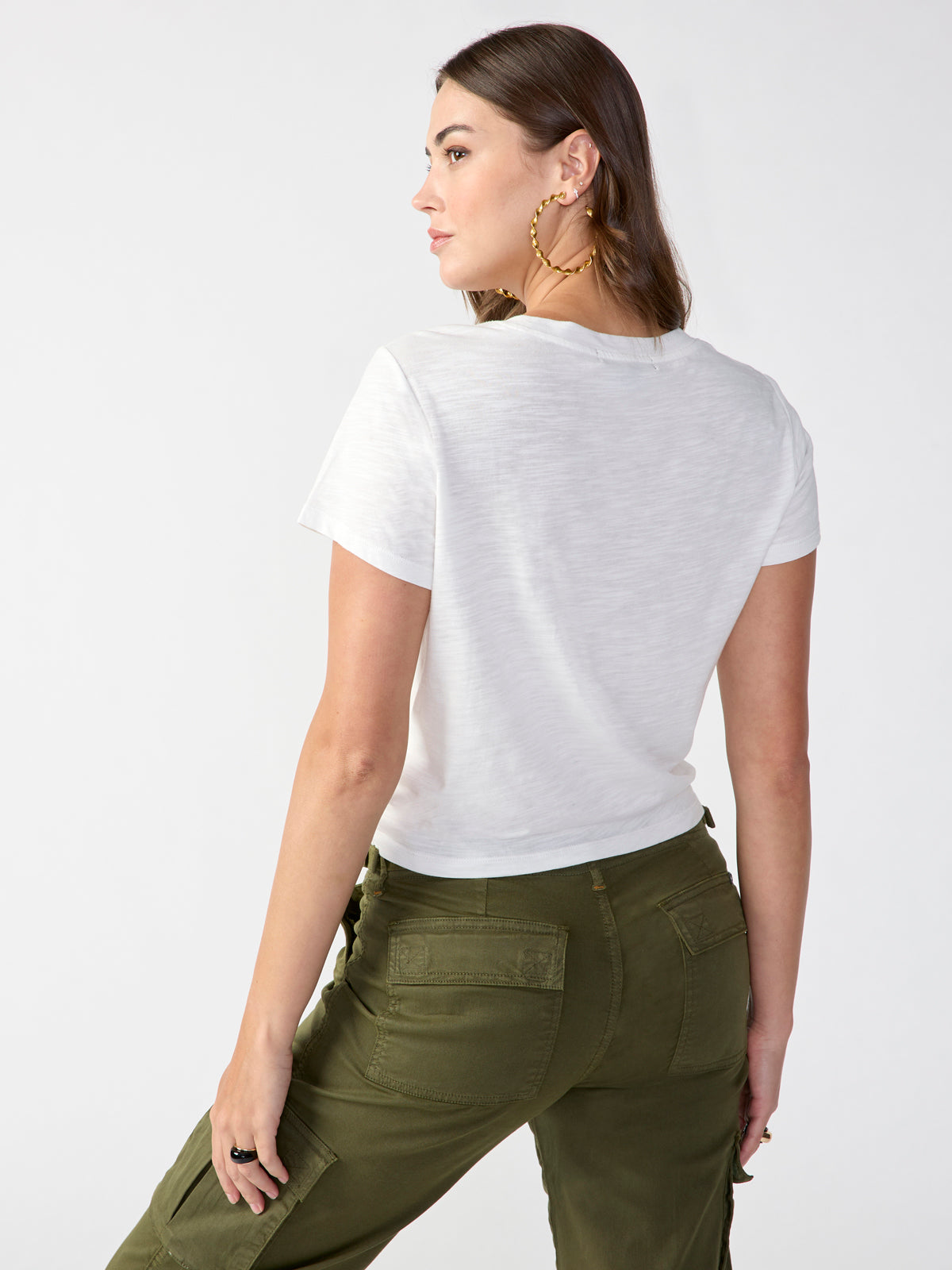 A person with long brown hair wearing large hoop earrings is posing with their back partially turned to the camera. They are dressed in a "Highstreet Tee White" from Sanctuary Clothing and olive green cargo pants, standing against a plain white background.