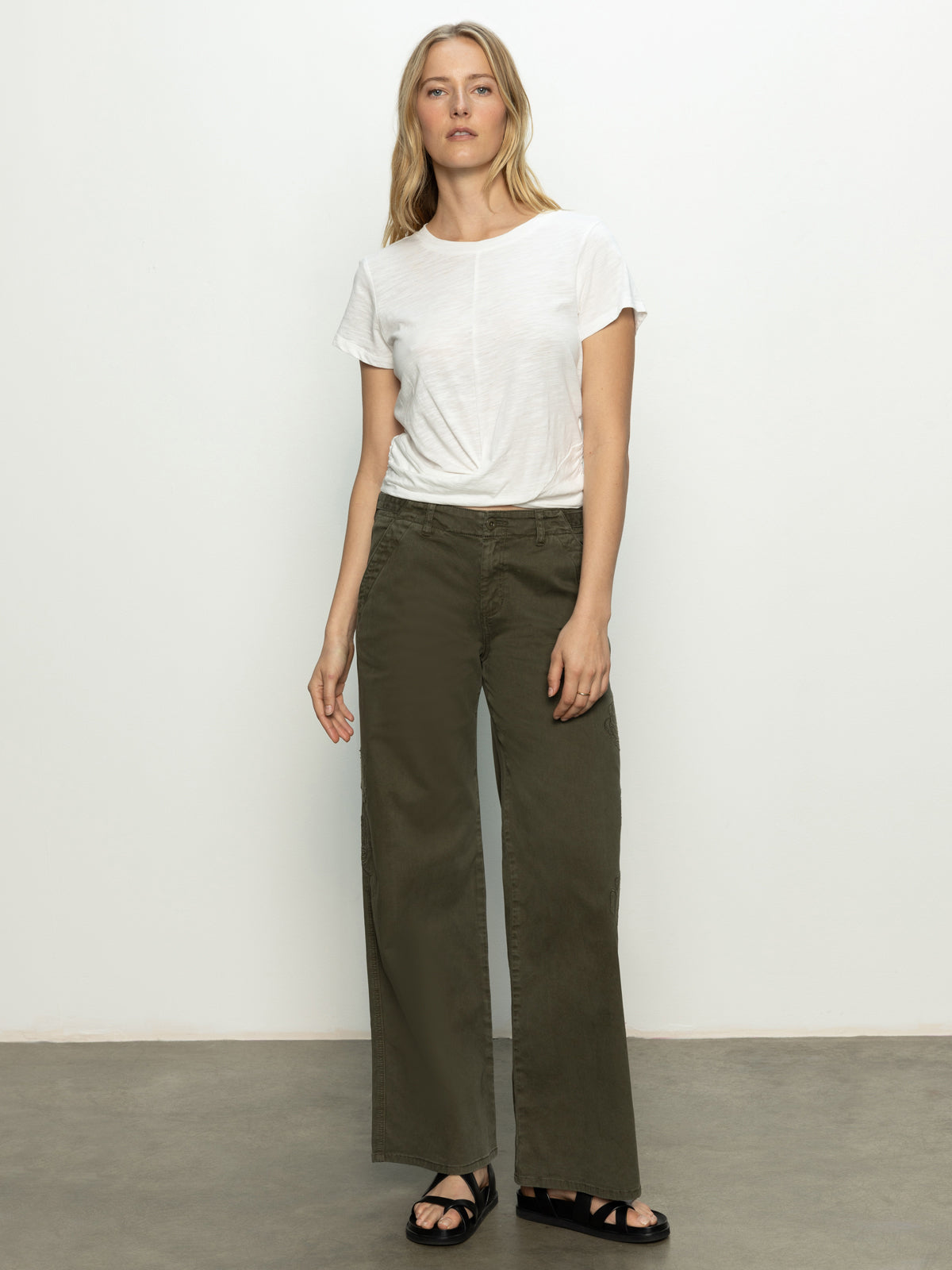 A person in a Sanctuary Clothing highstreet tee (white) and olive green wide-leg pants stands against a plain background. They have long, light hair, wear black sandals, and adopt a relaxed posture with one hand on their thigh.