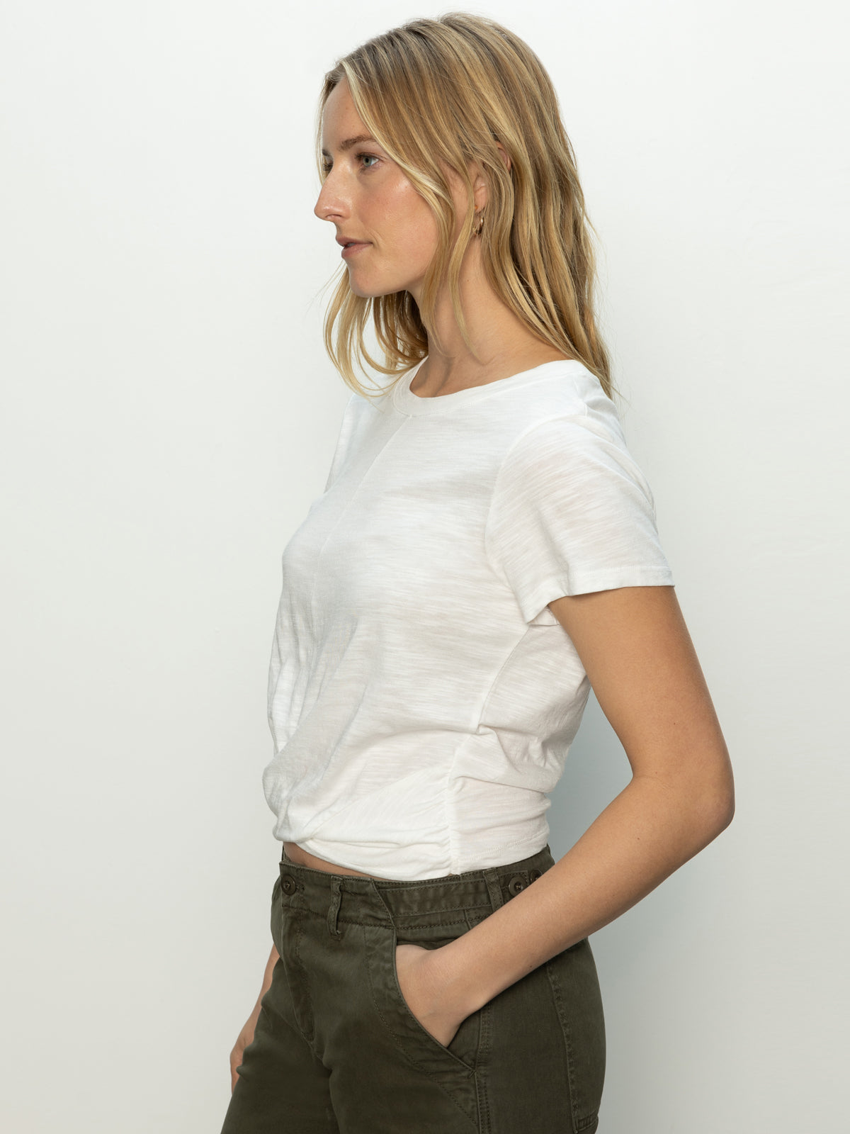 A person with long blonde hair stands in profile against a plain background, wearing Sanctuary Clothing's highstreet tee in white and olive green pants. They face forward with a neutral expression.