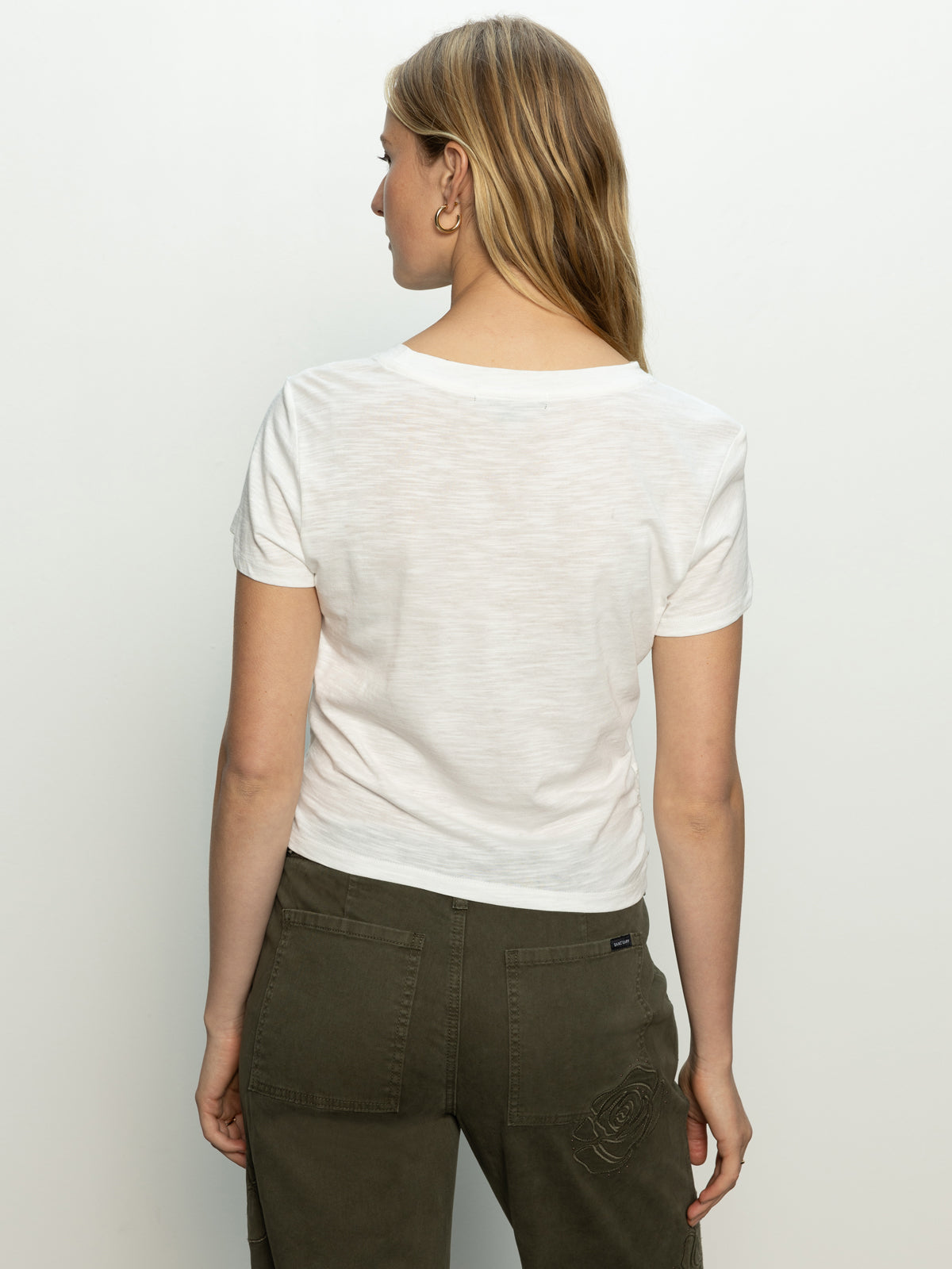 A person with long hair stands back to the camera, wearing a highstreet tee white by Sanctuary Clothing and olive green pants. The background is plain and light-colored.