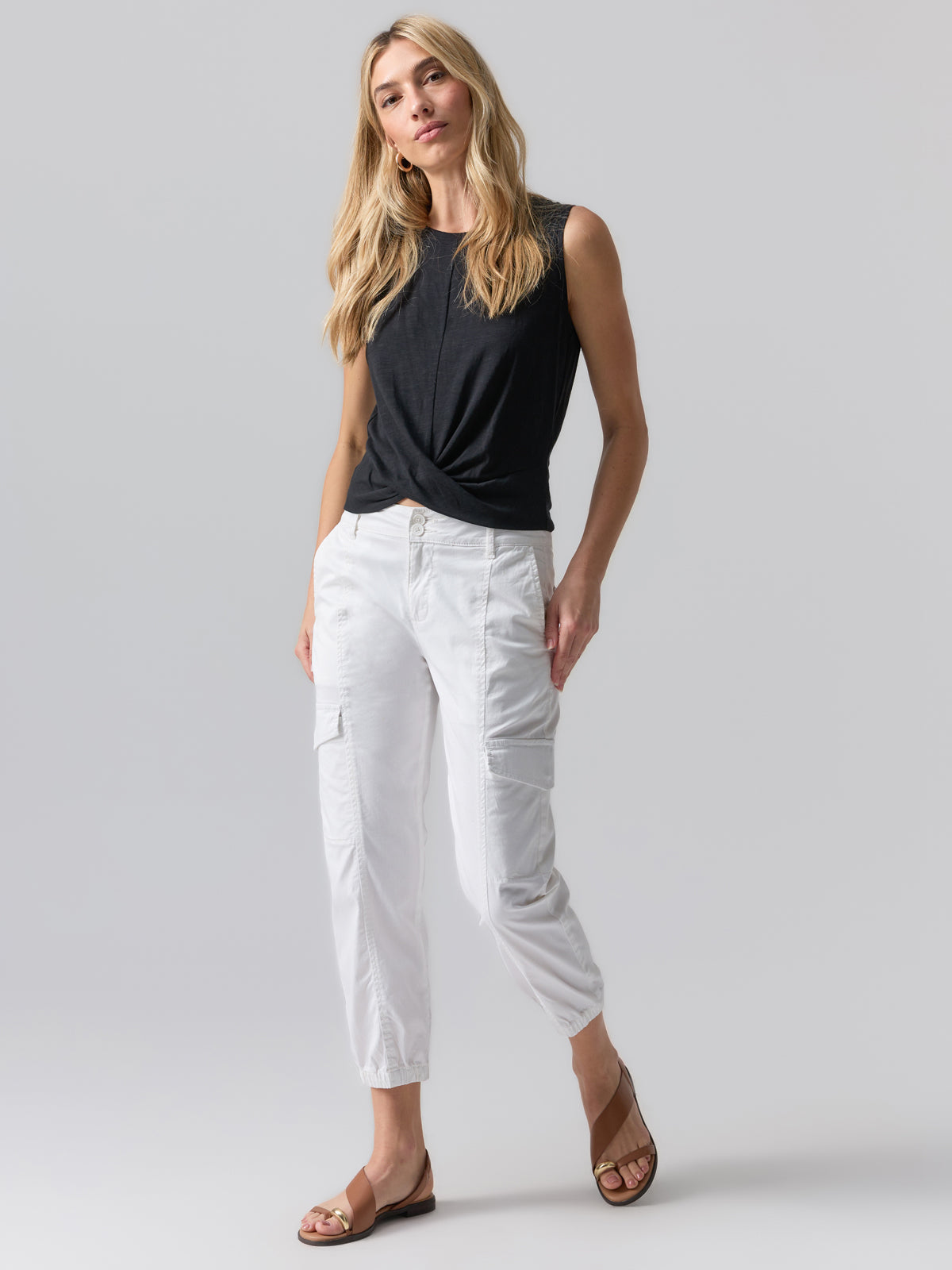 A woman stands against a plain background wearing the Twisted Tank Black by Sanctuary Clothing, white cargo pants, and brown sandals. She has long, wavy blonde hair and is posing with a relaxed, confident expression.