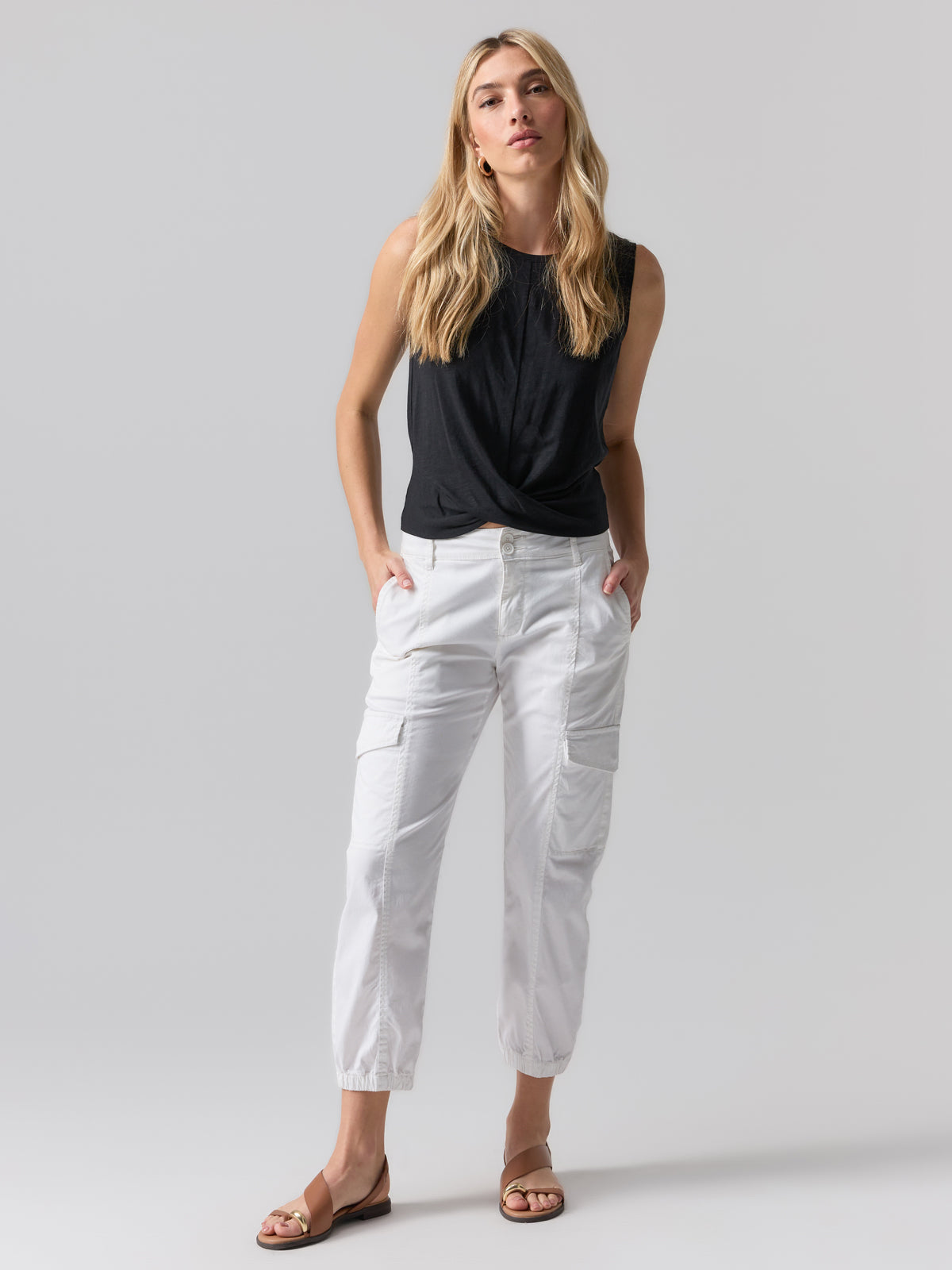 A woman with long blonde hair stands against a grey background, wearing the Twisted Tank Black by Sanctuary Clothing, white cargo pants, and brown sandals. She has her hands in her pockets and is gazing directly at the camera.