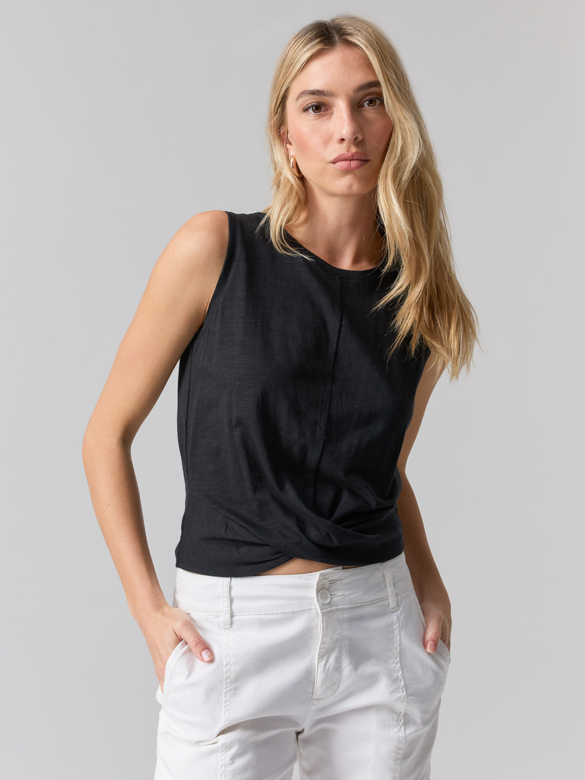 A person with long blonde hair is wearing the Twisted Tank Black by Sanctuary Clothing and white pants. They have their hands in their pockets and are looking directly at the camera with a neutral expression, set against a plain, light-colored background.